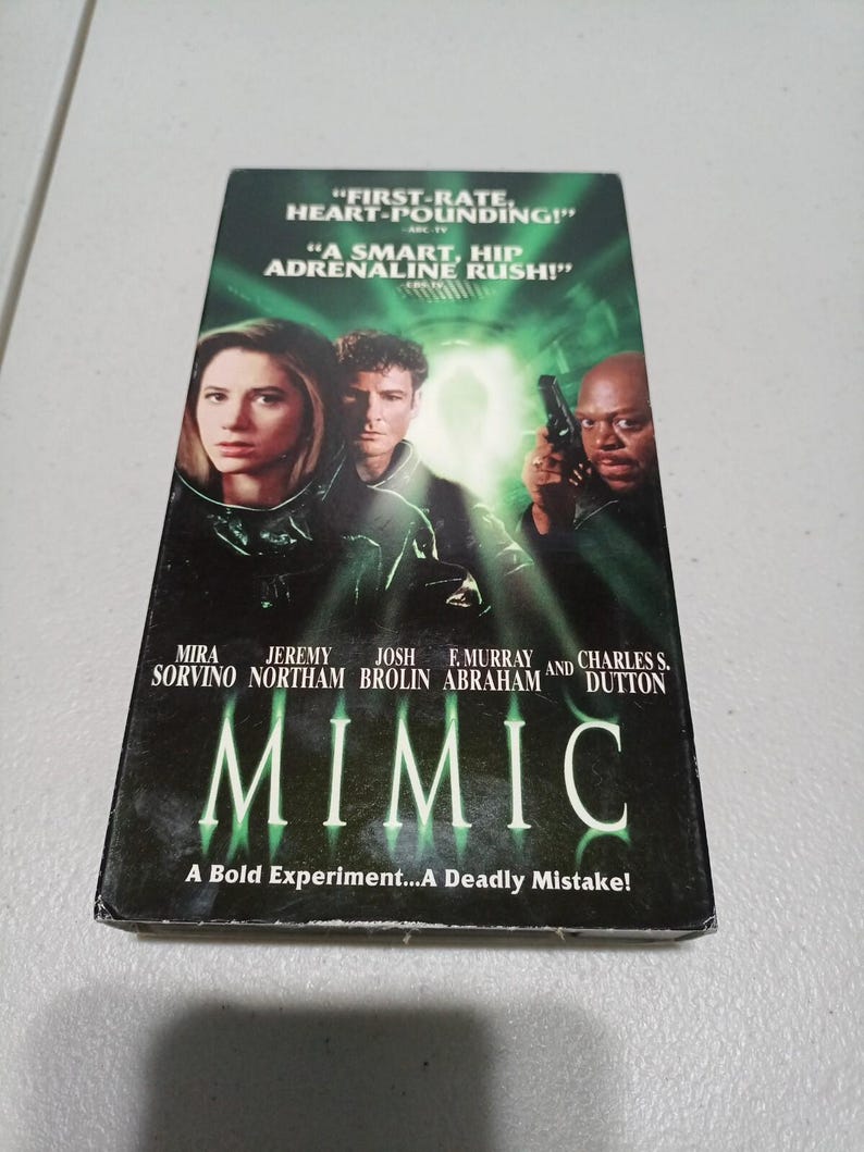 Może przedstawiać: Kaseta VHS z filmem Mimic z zielonkawą okładką przedstawiającą aktor&oacute;w: Mirę Sorvino, Jeremy'ego Northama, Josha Brolina, F. Murraya Abrahama i Charlesa S. Duttona. Tekst głosi First-Rate, Heart-Pounding, A Smart, Hip Adrenaline Rush!