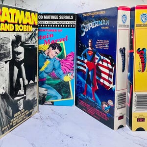 Może przedstawiać: Kolekcja starych kaset VHS z klasycznymi tytułami o superbohaterach i akcji. Kasety zawierają "Batman and Robin", "Adventures of Captain Marvel" i "TV's Best Adventures of Superman". Pudełka są kolorowe i zawierają oryginalne grafiki.