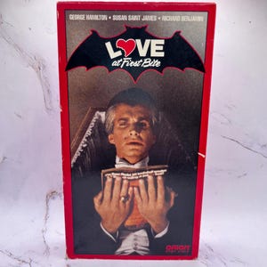 Może przedstawiać: Vintage pudełko po kasecie VHS z czerwoną obwódką zawiera tytuł filmu "LOVE at First Bite" z grafiką nietoperza. Okładka przedstawia mężczyznę w smokingu trzymającego książkę. Nazwiska aktorów są wymienione na górze.