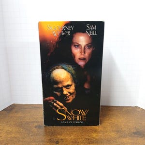 Może przedstawiać: Prostokątne pudełko na kasetę VHS z filmem Snow White: A Tale of Terror. Okładka przedstawia mroczny, nastrojowy obraz Sigourney Weaver i Sama Neilla nad zbliżeniem na bladą, starszą postać o złowrogim wyrazie twarzy i ostrych zębach.