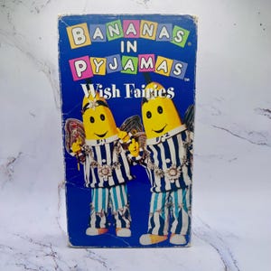 May include: A vintage VHS tape featuring the animated series "Bananas in Pyjamas: Wish Fairies." The cover is a vibrant blue, showcasing two banana characters in striped pajamas with fairy wings. The title text is in colorful, playful lettering.