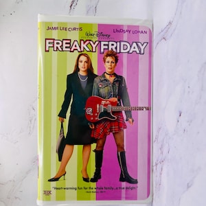 Puede incluir: Una cinta VHS de la película "Freaky Friday" con Jamie Lee Curtis y Lindsay Lohan. La portada tiene un fondo verde y rosa con el título de la película en letras grandes y en negrita. También es visible el eslogan de la película.