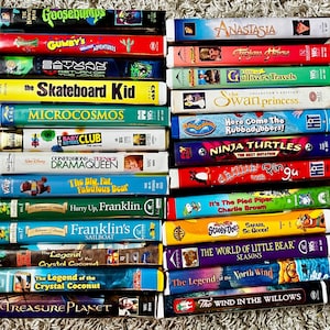 May include: A collection of VHS tapes featuring animated movies and children's shows. Titles include "Goosebumps," "Anastasia," "Ninja Turtles," and "Scooby-Doo." The tapes are stacked on a textured surface.