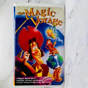 May include: VHS tape for "The Magic Voyage" animated film. The cover features a cartoon pirate, a fairy, and a worm-like creature on a globe. The title is in yellow and red, with text below stating it's a musical tale.