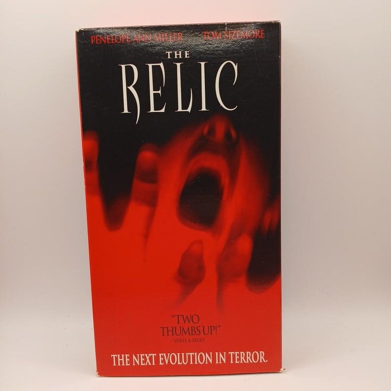 Może przedstawiać: Kaseta VHS z filmem The Relic w czarno-czerwonej kolorystyce. Okładka zawiera tytuł, nazwiska Penelope Ann Miller i Tom Sizemore, cytat Two Thumbs Up oraz napis The Next Evolution In Terror.