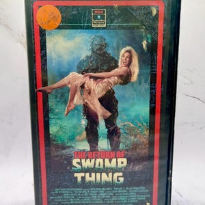 Puede incluir: Una cinta VHS vintage de la película "The Return of Swamp Thing". La portada presenta un monstruo sosteniendo a una mujer, con el título en letras verdes. La cinta tiene un borde rojo y el logotipo de RCA Columbia Pictures Home Video.