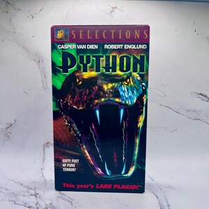 Puede incluir: Una cinta VHS de la película "Python" con Casper Van Dien y Robert Englund. La portada muestra un primer plano de la boca abierta de una serpiente con afilados colmillos. El texto incluye "Sixty Feet of Pure Terror!" y "This year's LAKE PLACID!"