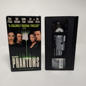 Może przedstawiać: Czarna kaseta VHS i jej kartonowe opakowanie do filmu Dean Koontz's Phantoms. Okładka przedstawia obsadę, tytuł dużymi białymi literami oraz tekst A CHILLINGLY ORIGINAL THRILLER. Czarna plastikowa kaseta posiada białą etykietę z napisami.