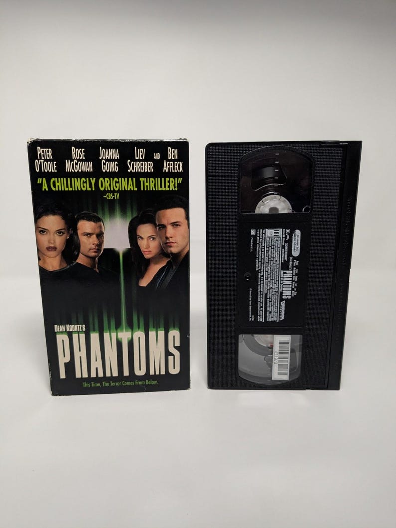 Może przedstawiać: Czarna kaseta VHS i jej kartonowe opakowanie do filmu Dean Koontz's Phantoms. Okładka przedstawia obsadę, tytuł dużymi białymi literami oraz tekst A CHILLINGLY ORIGINAL THRILLER. Czarna plastikowa kaseta posiada białą etykietę z napisami.