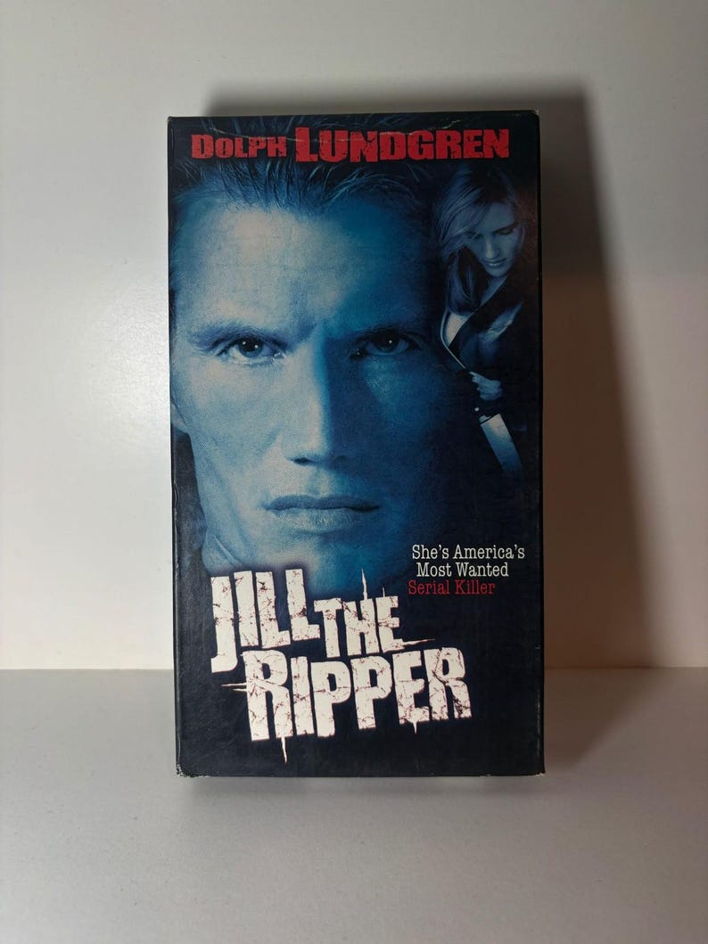 Może przedstawiać: Kaseta VHS z filmem Jill the Ripper z Dolphem Lundgrenem w roli gł&oacute;wnej. Okładka przedstawia duży, niebieskawy portret mężczyzny, mniejszy obraz kobiety trzymającej n&oacute;ż oraz tekst Jill the Ripper i She's America's Most Wanted Serial Killer.