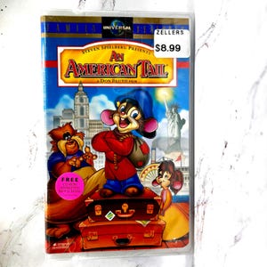 Może przedstawiać: Etui DVD z filmem animowanym An American Tail, przedstawiające Fievela Mousekewitza stojącego na dwóch ułożonych w stos brązowych walizkach. Okładka zawiera panoramę miasta, Statuę Wolności oraz różową naklejkę z napisem FREE CD-ROM ANIMATED MOVIE BOOK.