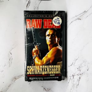 Peut inclure: Une cassette VHS du film "Raw Deal" avec Arnold Schwarzenegger. La couverture présente le titre en rouge, l'acteur tenant une arme et le texte "Collector's Edition". Une étiquette de prix est dans le coin supérieur droit.