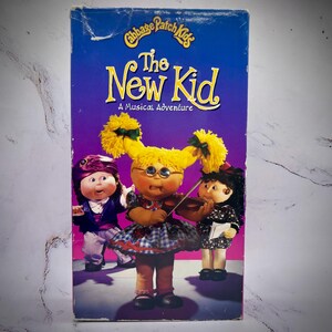 May include: A vintage VHS tape for "Cabbage Patch Kids: The New Kid, A Musical Adventure." The cover features three Cabbage Patch Kids dolls, one playing a violin. The title is in yellow, with a blue and purple background.