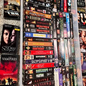 Może przedstawiać: Kolekcja licznych kaset VHS ułożonych w przezroczystym plastikowym pojemniku do przechowywania oraz rozrzuconych wok&oacute;ł niego. Grzbiety kaset prezentują r&oacute;żne tytuły film&oacute;w, w tym The Car, Snow White, Vampires, Alien Visitor, The Skulls i Panic Room.