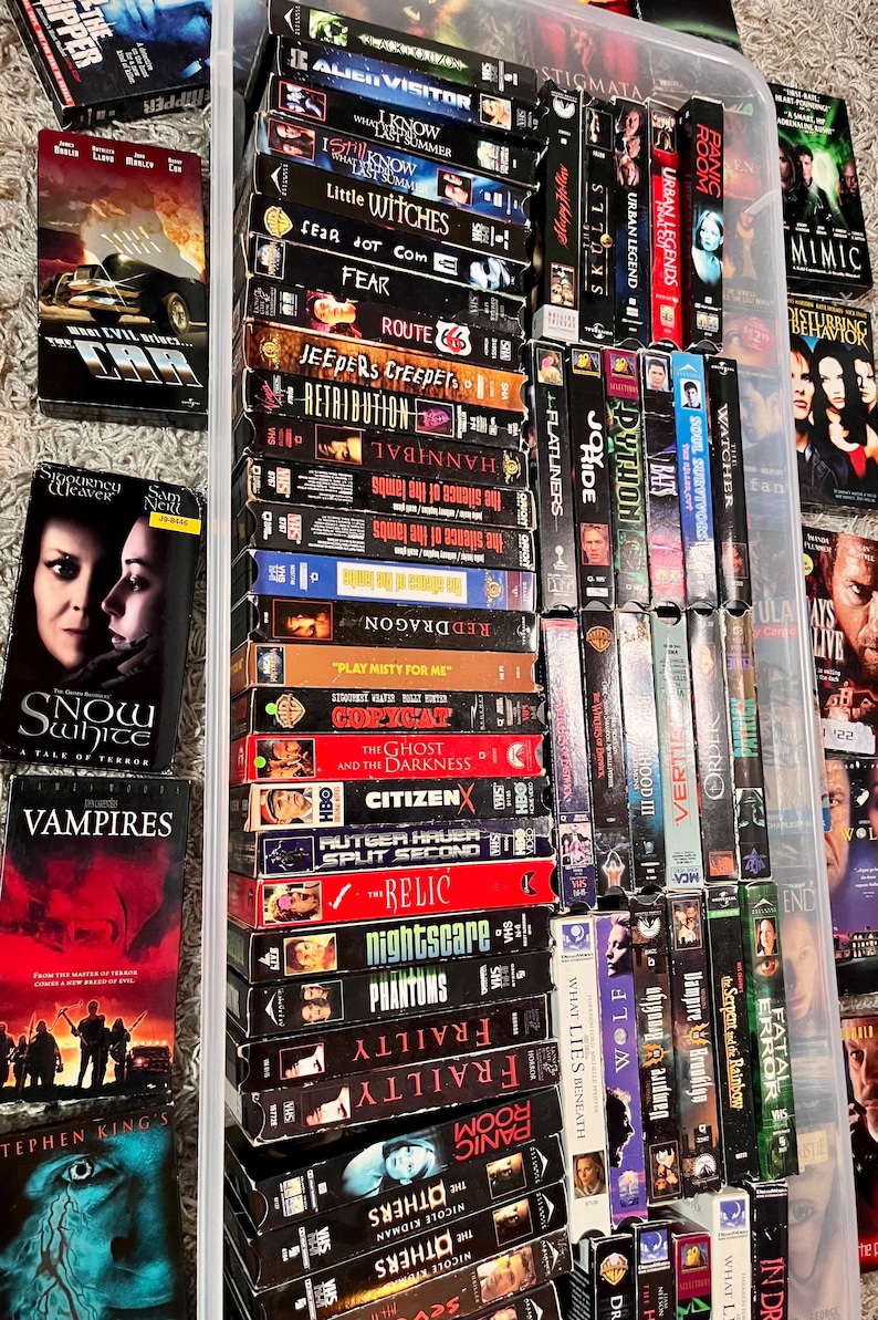 Może przedstawiać: Kolekcja licznych kaset VHS ułożonych w przezroczystym plastikowym pojemniku do przechowywania oraz rozrzuconych wok&oacute;ł niego. Grzbiety kaset prezentują r&oacute;żne tytuły film&oacute;w, w tym The Car, Snow White, Vampires, Alien Visitor, The Skulls i Panic Room.