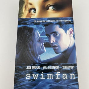Może przedstawiać: Kaseta VHS z filmem Swimfan, przedstawiająca zbliżenie niebieskich oczu u g&oacute;ry, mężczyznę i kobietę patrzących na siebie w środku oraz tło z niebieskimi falami wody u dołu z napisem His biggest fan just became his worst nightmare.
