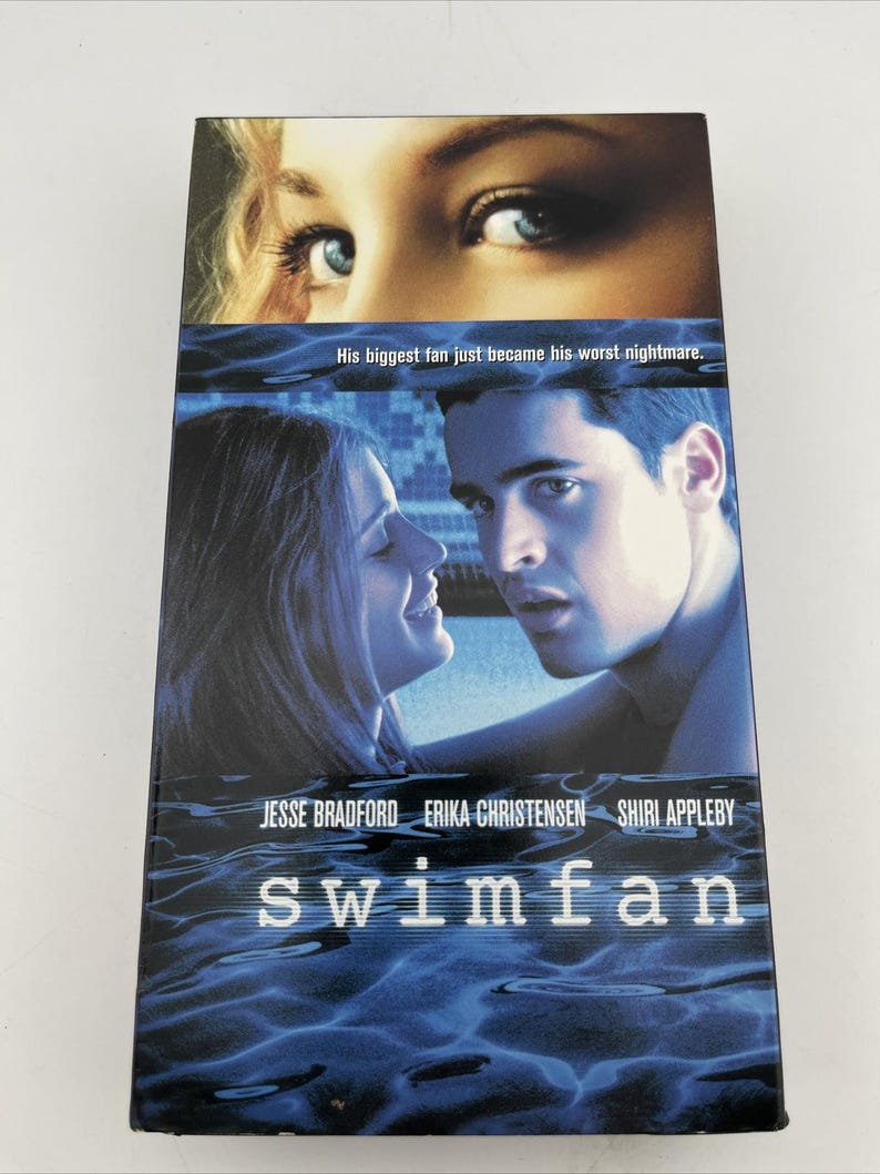 Może przedstawiać: Kaseta VHS z filmem Swimfan, przedstawiająca zbliżenie niebieskich oczu u g&oacute;ry, mężczyznę i kobietę patrzących na siebie w środku oraz tło z niebieskimi falami wody u dołu z napisem His biggest fan just became his worst nightmare.