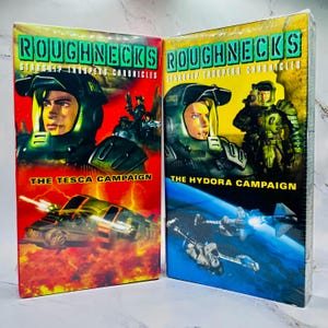 May include: Two VHS tapes of "Roughnecks: Starship Troopers Chronicles". The first tape, "The Tesca Campaign", has a red background with a spaceship. The second tape, "The Hydora Campaign", has a blue background with spaceships. Both tapes feature a soldier in a helmet.