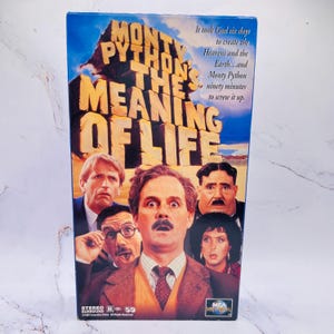 May include: A VHS video cassette of "Monty Python's The Meaning of Life." The cover shows a group of actors with shocked faces, the film's title in bold letters, and the quote, "It took God six days to create the Heavens and the Earth...and Monty Python ninety minutes to screw it up."