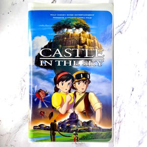 May include: A VHS video tape for the animated film "Castle in the Sky." The cover shows an illustration of two characters, a floating castle, and the film's title. The tape is in a plastic case.