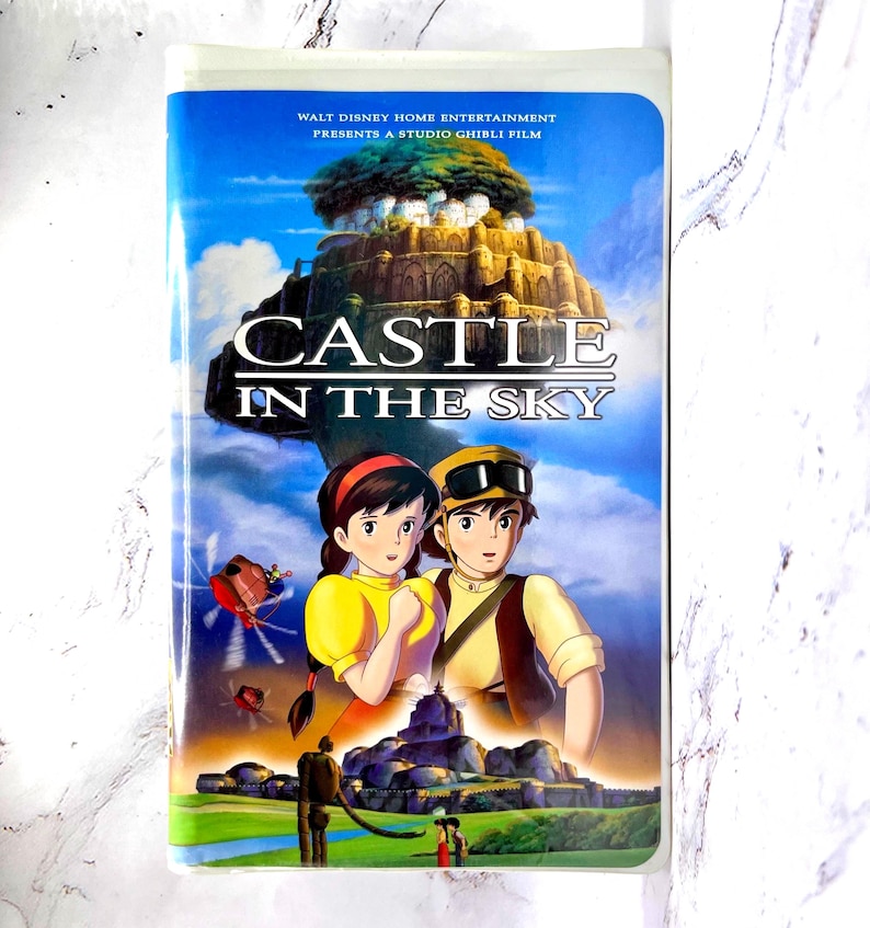 May include: A VHS video tape for the animated film "Castle in the Sky." The cover shows an illustration of two characters, a floating castle, and the film's title. The tape is in a plastic case.