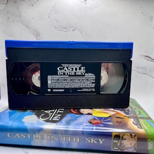 May include: A black VHS video cassette labelled "CASTLE IN THE SKY" with a blue top. The tape is on top of a VHS case with the same title and colourful artwork. The Disney logo is visible.