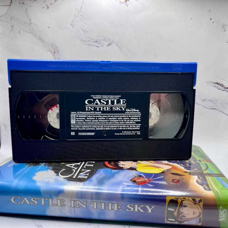 May include: A black VHS video cassette labelled "CASTLE IN THE SKY" with a blue top. The tape is on top of a VHS case with the same title and colourful artwork. The Disney logo is visible.