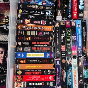 Może przedstawiać: Kolekcja licznych kaset VHS ułożonych w przezroczystym plastikowym pojemniku. Grzbiety kaset są widoczne, prezentując r&oacute;żne tytuły film&oacute;w, takie jak The Silence of the Lambs, Red Dragon, Copycat i The Relic, wraz z logotypami studi&oacute;w filmowych.