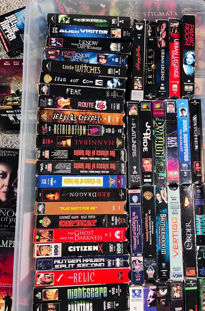 Może przedstawiać: Kolekcja licznych kaset VHS ułożonych w przezroczystym plastikowym pojemniku. Grzbiety kaset są widoczne, prezentując r&oacute;żne tytuły film&oacute;w, takie jak The Silence of the Lambs, Red Dragon, Copycat i The Relic, wraz z logotypami studi&oacute;w filmowych.