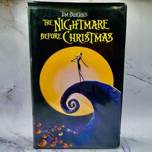 Peut inclure: Une cassette VHS noire de "L'Étrange Noël de Monsieur Jack" de Tim Burton. La couverture présente Jack Skellington sur une colline bleue tourbillonnante devant une lune jaune. Le titre est en lettres jaunes et blanches.
