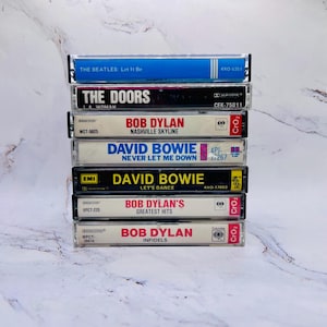 May include: A stack of vintage cassette tapes featuring iconic artists like The Beatles, The Doors, Bob Dylan, and David Bowie. The tapes have colorful labels with artist names and album titles. The stack is on a white surface.