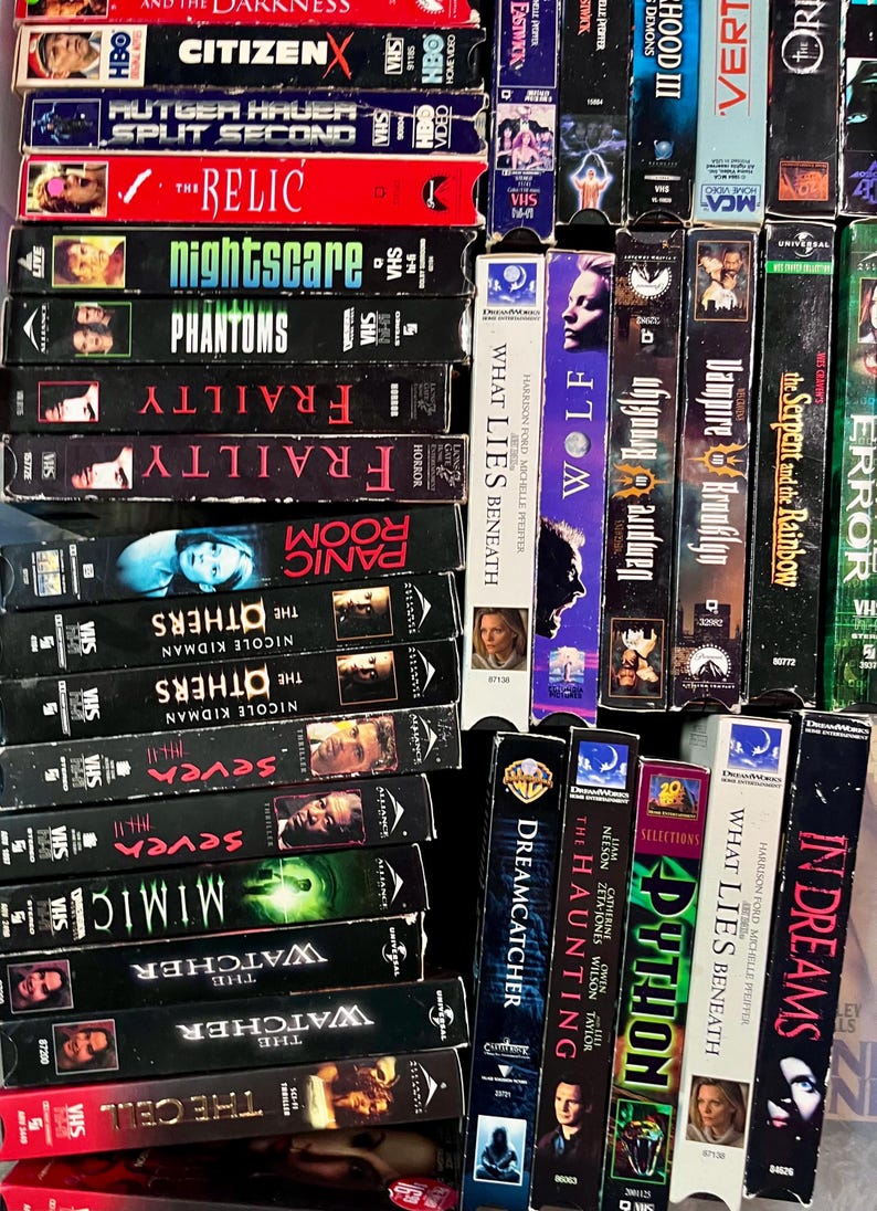 Może przedstawiać: Kolekcja kaset VHS ułożonych w siatkę, zawierająca tytuły takie jak The Relic, Nightscare, Phantoms, Frailty, Panic Room, The Others, Seven, Mimic, The Watcher, The Cell, What Lies Beneath, Wolf, Vampire in Brooklyn oraz In Dreams.