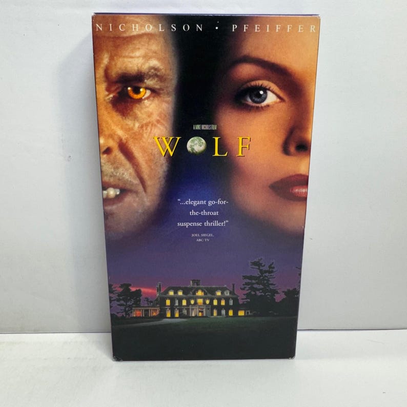 Może przedstawiać: Kaseta VHS z filmem Wolf, przedstawiająca twarze Jacka Nicholsona i Michelle Pfeiffer. Tytuł Wolf jest wydrukowany na ż&oacute;łto z grafiką księżyca zamiast litery O. Poniżej znajduje się dom o zmierzchu wraz z cytatem autorstwa Joela Siegela.