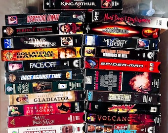 May include: A collection of VHS tapes featuring movie titles like "King Arthur," "Vertical Limit," "The Perfect Storm," and "Gladiator." The tapes are stacked, showcasing various movie covers with actors and film titles.