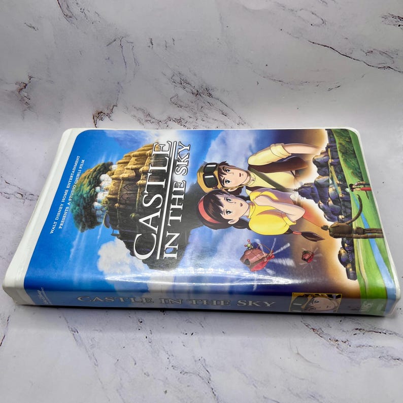 May include: A VHS tape of the animated film "Castle in the Sky." The cover features the title in white, with a blue sky background and illustrations of characters and a floating island. The tape is white.