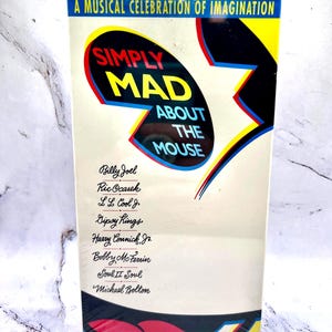 Może przedstawiać: Kaseta VHS z napisem "SIMPLY MAD ABOUT THE MOUSE" w kolorowym designie. Na kasecie znajduje się lista artystów muzycznych, w tym Billy Joel, Ric Ocasek i Michael Bolton. Na górze kasety widnieje napis "A MUSICAL CELEBRATION OF IMAGINATION."