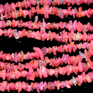 May include: Close-up of several strands of pink gemstone chips, possibly opal or tourmaline, against a black backdrop. The chips, in varying sizes and shapes, display a range of pink shades. The beads are strung, indicating their use in jewellery.