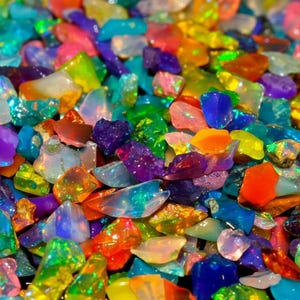 May include: A close-up of a pile of colorful, iridescent gemstones. The gemstones are a mix of colors, including red, orange, yellow, green, blue, and purple. The stones are irregularly shaped and have a glossy finish.