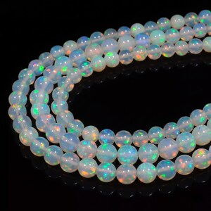 May include: A strand of round, translucent white opal beads featuring a play-of-color effect with flashes of green, blue, and orange light. The beads are arranged in a curved, multi-layered formation against a solid black background.