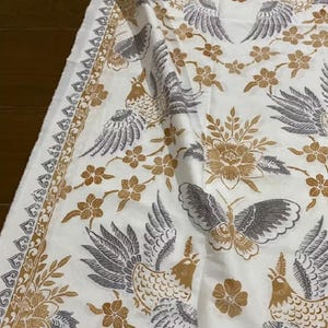 Puede incluir: Tela blanca con un patrón repetido de aves estilizadas y elementos florales en tonos grises y dorados. La tela tiene un borde detallado con un diseño repetido. La estética general recuerda a los diseños textiles tradicionales.