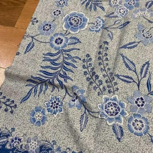 Könnte beinhalten: Ein Stofftextil mit hellgrauem Hintergrund und einem dichten, komplizierten Muster aus blauen und weißen Blumenmotiven, einschließlich stilisierter Blüten und blättriger Ranken. Das Design enthält einen markanten Rand mit einem dunkelblauen Punktmuster.