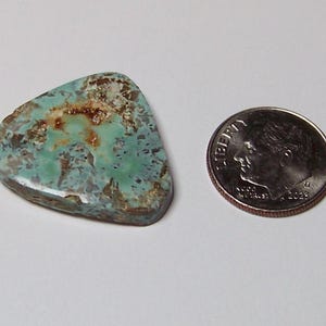 May include: A triangular turquoise gemstone with a polished surface featuring a mix of light blue, green, and brown matrix patterns. The stone is positioned next to a standard United States dime for scale, showing its relative size in this view.