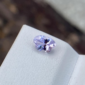 May include: A light purple, oval-cut gemstone. The gem is displayed on a white surface, with a blurred background. The facets of the gemstone catch the light, creating a sparkling effect. This gem could be used in jewellery.