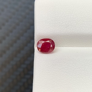 Può includere: Una vibrante gemma di rubino a taglio ovale. La gemma rosso intenso è esposta su una superficie bianca, mostrando le sue sfaccettature lucide e il suo colore ricco. La gemma è il punto focale, evidenziando la sua bellezza naturale.