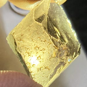 Puede incluir: Primer plano de un objeto dorado de forma irregular, posiblemente una gema o un trozo de metal. El objeto tiene una superficie texturizada con imperfecciones visibles y una cualidad reflectante.