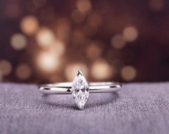 Pode incluir: Um anel de prata com um diamante lapidação marquise. O diamante é o ponto focal, cravado em uma banda fina. O anel é exibido em uma superfície cinza com um fundo desfocado de luzes quentes fora de foco.