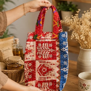 May include: A rectangular quilted tote bag featuring a patchwork design with red, cream, and blue floral and geometric patterns. The bag has two matching fabric handles and visible white running stitch embroidery throughout the quilted surface.