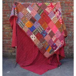 Könnte beinhalten: Ein Patchwork-Quilt mit einer lebendigen Mischung aus Farben und Mustern, darunter Quadrate aus roten, rosa, orangefarbenen, blauen und violetten Stoffen. Der Quilt ist über einer einfarbigen roten Decke drapiert. Der Hintergrund ist eine Backsteinmauer.