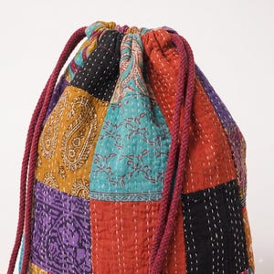 Op de afbeelding: Een buidel met trekkoord gemaakt van veelkleurige patchworkstof met diverse patronen, waaronder paisley- en bloemmotieven. De tas is geconstrueerd met zichtbare witte stikseldetails en bevat twee dikke, bordeauxrode gevlochten koorden.