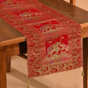 Puede incluir: Camino de mesa rojo con diseños dorados de elefantes y flores. El camino de mesa tiene forma rectangular, con un borde detallado y borlas en los extremos. Los elefantes están representados en un estilo ornamentado.